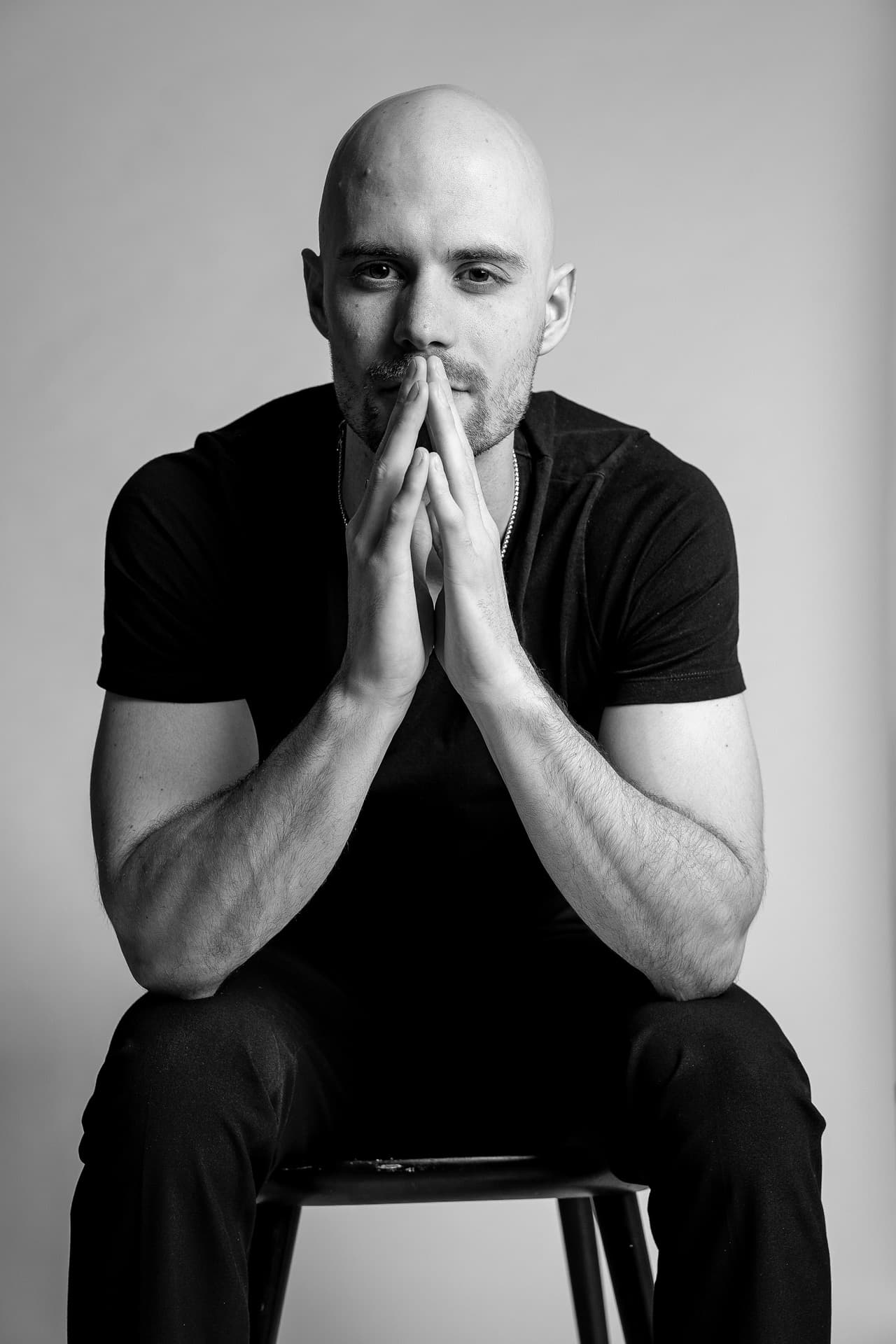 Black-and-white photo of Andrei Potekhin seated on a stool, hands pressed together in front of his face.